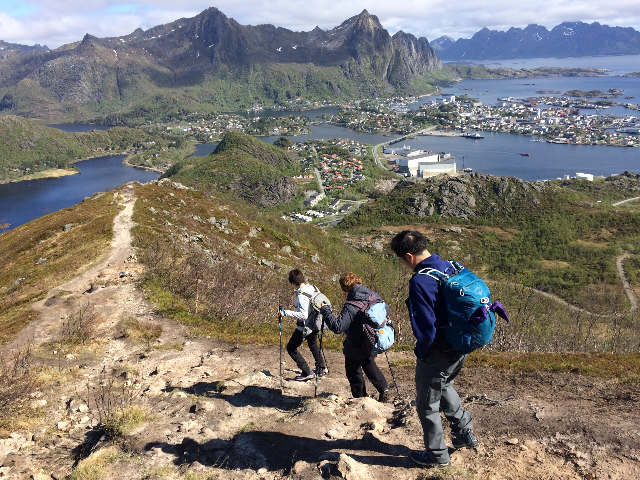 Three hikers descending a mountain trail with a panoramic view of Svolvær, Lofoten and surrounding fjords and peaks. Hiking in Lofoten guide